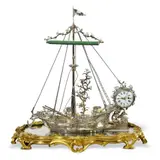 François-Thomas Germain, For the seal bearer of King Louis XV: The ecritoire of Jean-Baptiste de Machault d'Arnouville, Silver, porcelain, mirror pane, bronze, gilding. An ecritoire formed as a detailed model of a sailing boat with mast and rigging placed upon a glass mirror pane resembling still water and mounted on an ormolu base with raised rococo décor. The inkwell and pounce box concealed within the two barrels on the deck and with small lidded compartments in the fish traps at the stern of the boat. The raised anchor can be seen at the bow, and below the stern the rudder formed as a finely chased dolphin. On the port side we see tiny fish and crustaceans caught in a finely knotted silver net, the starboard side fitted with a candlestick with two nozzles. When the fabric sail is unfolded, it forms a screen to protect from the light. A large cylindrical clock with a white enamel dial over the stern of the boat entwined by a climbing rose with polychrome Vincennes porcelain flowers that extends up towards the rigging. H 41, W 39.8, D