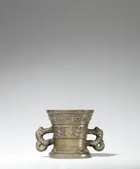 Werkstatt Hachmann, A small Hachmann mortar with an inscription, Golden brown cast bronze with natural patina. The display side decorated with a vacant coat-of-arms supported by winged demi-figures. Two broad banderoles above the base and below the rim inscribed in Latin capital letters: “ANNO DOMIN M VC LXXVI” and “N DEO SPES MEA”. With zoomorphic handles. Dents to the rim. H 11.3, D