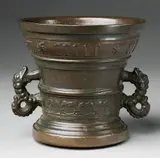 A dated Hachmann mortar, A dated Hachmann mortar, Cast bronze, the golden brown patination presumably re-applied and with traces of green corrosion. A fleur-de-lys band surrounding the lower border beneath a vacant shield supported by two winged half-figures. A broad banderole around the rim inscribed "anno domini MCCCCCXLVI". The handles formed as two-headed dragons. H 13.9, D