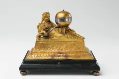Hanns Buschmann, An Augsburg figural clock "Urania", Gilt copper and bronze, engraved dial and silver hands, ebonised wood base and later red velvet to the interior. One day movement with hourly striking to a bell. Designed as a figure of Urania, the muse of astrology, recumbent upon a moulded gilt copper plinth upon a contrasting ebonised wood base. The sceptre in her right hand forms the hour hand of the clock, pointing towards the hour indices on the equatorial band of the globe on a column. Engraved to the upper right of the plate "Hannß Buschmann A". H 16.3, W 20, D