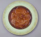 Crown Lynn Wharetana Ware, 'Hei Tiki' wall plate, with a brown glazed centre and a green glazed border (with hanging wire through suspension holes to foorim)