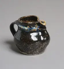 Small Fatso jug by Barry Brickell, tenmoku glazed stoneware with blue jun glaze highlights to the rim, typical pinched rim with stylized characterful beak , sold Feb 2026
