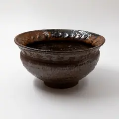 Sculpted bowl by Barry Brickell, grogged clay stoneware with tenmoku glaze, relief cross pattern around the flaring rim (minor fritting to the rim), sold Feb 2026