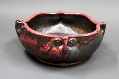 Stoneware bowl by Graeme Storm, shaped with the rim pinched with four mask forms, manganese glaze and copper red highlights, offered May 2025