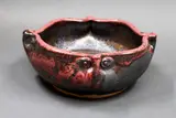 Stoneware bowl by Graeme Storm, shaped with the rim pinched with four mask forms, manganese glaze and copper red highlights, offered May 2025