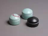 Three porcelain lidded boxes by John Dawson (New Zealand & Uk), one in matt black glaze, one in pale celadon glaze, the third in pale celadon and red gestural decoration to the lid, offered May 2024