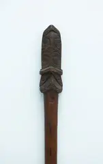 Taiaha, Fighting staff, carved from medium brown wood, slender plain shaft tapering into a narrow rounded blade. Carved upoko and arero with double raupongo spiral pattern, plain eyes.