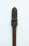 Taiaha, Fighting staff, carved from medium brown wood, slender plain shaft tapering into a narrow rounded blade. Carved upoko and arero with double raupongo spiral pattern, plain eyes.