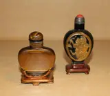 Two Chinese tiger's eye snuff bottles, 19th/20th Century, the first with tigers eye stopper, the second carved with figural panels, hardstone and metal stopper, both with wood stands