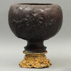 Macetero, JAPÓN, CA, 1900, Bronce, patinado en color marrón; decorado con relieves a manera de aves en enramada y representación de deidad con dragones; se incluye una base de bronce dorado con relieves de águilas y figuras florales (matrimonio)