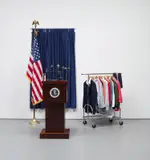 Mark Dion, Mess Conference, 2004, wooden podium, microphones, curtain rail, curtains, flags, flag pole, framed black and white photographs, books, glass, badges, costume jewellery, hair accessories, glasses, hair brushes, scarves, shirts, ties, jackets, skirts, coat hangers, clothes rail and card-mounted pie chart