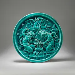 Majolica wall charger, early 20th Century by Clement Massier, Earthenware, turquoise-blue glaze, depicting in high relief three putto, forging arrows in a fire and at an anvil, sold Dec 2023