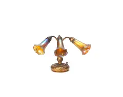 A Three-Light Lily Piano Lamp by Tiffany Studios, circa 1910, gilded bronze, the base cast with foliate detail, the Favrile glass shades in a golden and petrol blue iridescent glass, offered Jun 2019