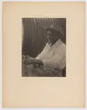 THE BASKETWEAVER SOUTH CAROLINA GULLAH PHOTOGRAPHIC PORTRAIT by Doris Ulmann, platinum print depicting an African American man smoking a pipe and weaving a basket, sold Dec 2023